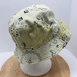 May include: A cream and olive green bucket hat with a paisley pattern. The hat features a patchwork design with a white paisley print and black accents. The brim is lined with a matching paisley fabric.