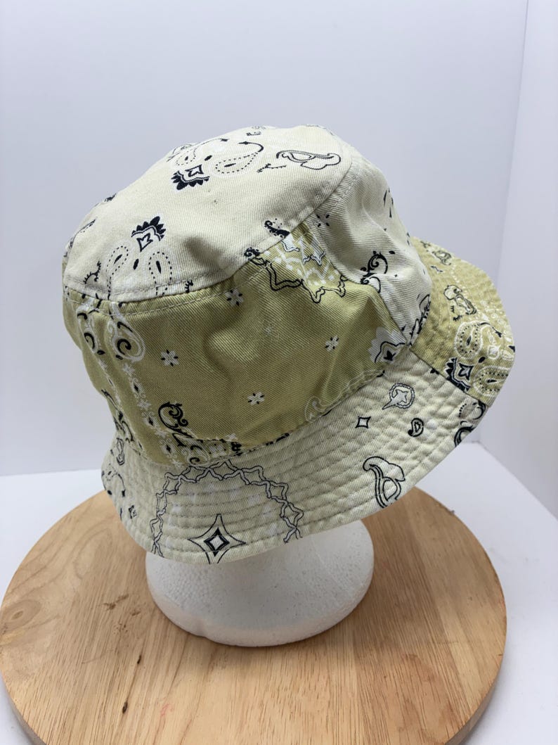 May include: A cream and olive green bucket hat with a paisley pattern. The hat features a patchwork design with a white paisley print and black accents. The brim is lined with a matching paisley fabric.