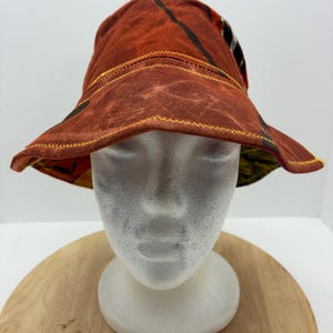 May include: A burnt orange bucket hat with a patterned design. The hat features a wide brim and is constructed with visible yellow stitching. The hat is displayed on a white mannequin head, set against a neutral background.