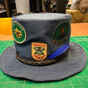 May include: A blue denim top hat adorned with three embroidered patches and a blue feather. The patches include a green Patrol Leader emblem, a Pinewood Racer car graphic, and a circular green badge. A dark brown leather band with metal studs encircles the base.