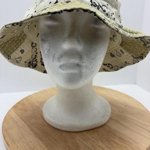 May include: Cream-colored bucket hat with a black paisley pattern. The hat is reversible, with a light green paisley pattern on the underside. The hat is displayed on a white mannequin head.
