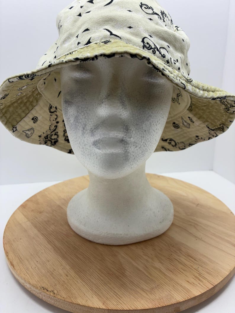 May include: Cream-colored bucket hat with a black paisley pattern. The hat is reversible, with a light green paisley pattern on the underside. The hat is displayed on a white mannequin head.