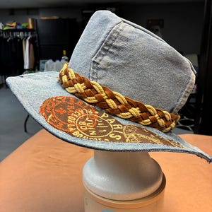 May include: A denim hat with a brown woven band and a decorative brim. The brim features a printed design with the word "Paris". The hat is displayed on a white mannequin head.