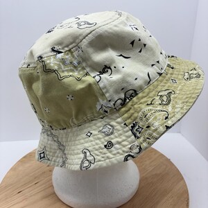 May include: A patchwork bucket hat in shades of cream and olive green. The hat features a paisley pattern in black and white. The hat is made of a soft, possibly cotton, material.