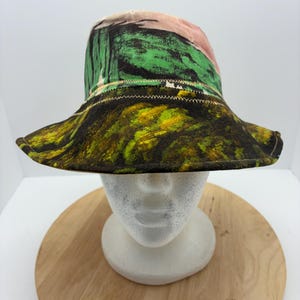 May include: A colorful bucket hat with a wide brim. The hat features a vibrant mix of colors and patterns, including green, yellow, black, and pink. The brim has a marbled effect, and the crown has a painted design.