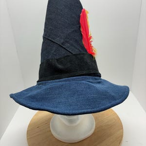 May include: A denim hat with a wide brim and a tall, pointed crown. The hat features a black band and is adorned with a red and yellow feather. The hat is displayed on a white mannequin head and a wooden surface.