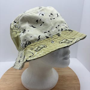 May include: A reversible bucket hat with a paisley pattern. The hat features a light green and off-white color scheme. The brim and the top of the hat have a light green paisley pattern, while the rest of the hat is off-white with black paisley designs.