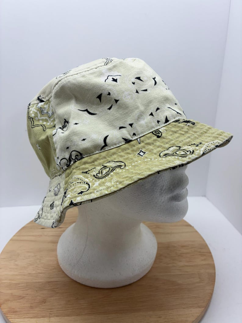 May include: A reversible bucket hat with a paisley pattern. The hat features a light green and off-white color scheme. The brim and the top of the hat have a light green paisley pattern, while the rest of the hat is off-white with black paisley designs.