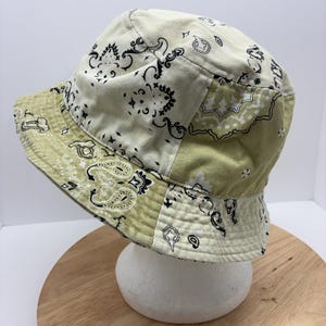 May include: A patchwork bucket hat with a paisley pattern in cream and olive green. The hat features a classic brim and a soft, casual style. The hat is displayed on a white foam head.