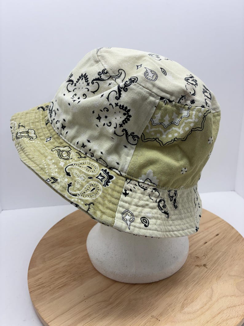 May include: A patchwork bucket hat with a paisley pattern in cream and olive green. The hat features a classic brim and a soft, casual style. The hat is displayed on a white foam head.