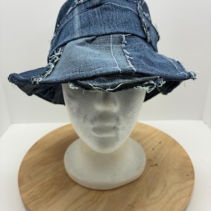 May include: A blue denim bucket hat with a frayed brim and patchwork design. The hat is made from recycled denim and features visible stitching. The hat is displayed on a white mannequin head.