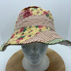 May include: A patchwork bucket hat with a floral and gingham pattern. The hat features a mix of fabrics in shades of yellow, red, and green. The brim is lined with a green and white trim. The hat is displayed on a white mannequin head.