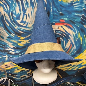 May include: A blue denim pointed hat with a wide brim and a textured tan woven band around the base. The hat is displayed on a white mannequin head, featuring a dark brown feather accent tucked into the side of the band near the crown.