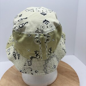May include: A two-tone bucket hat with a paisley pattern. The hat features a light green brim and a cream-colored crown, both adorned with black and white paisley designs. The hat is displayed on a white form against a light background.