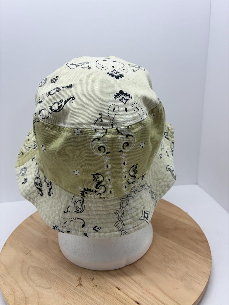 May include: A two-tone bucket hat with a paisley pattern. The hat features a light green brim and a cream-colored crown, both adorned with black and white paisley designs. The hat is displayed on a white form against a light background.