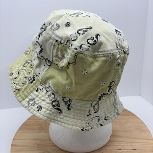 May include: A patchwork bucket hat in shades of cream and olive green, featuring a paisley pattern in black. The hat is made of fabric and has a rounded crown and a wide brim. The hat is displayed on a white foam head.