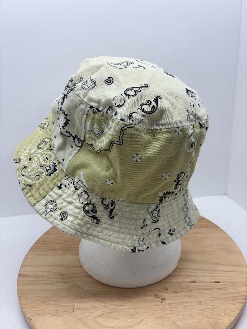 May include: A patchwork bucket hat in shades of cream and olive green, featuring a paisley pattern in black. The hat is made of fabric and has a rounded crown and a wide brim. The hat is displayed on a white foam head.