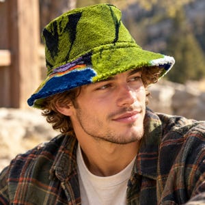 May include: A fuzzy bucket hat featuring a vibrant green base with abstract patterns in black, blue, purple, and orange. The hat has a textured, plush surface and a wide, downward-sloping brim that provides a distinct silhouette.