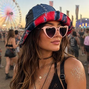 May include: A bucket hat featuring a patchwork design of dark blue denim and a red and black tie-dye pattern with frayed edges, paired with light pink heart-shaped sunglasses that have dark gradient lenses and a thin plastic frame.