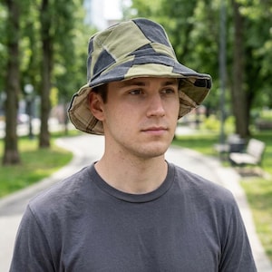 May include: A patchwork bucket hat in shades of green, black, and gray. The hat has a wide brim and a patterned design. The person in the image is wearing a gray t-shirt.