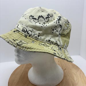 May include: A light green and cream-colored bucket hat with a paisley pattern. The hat features a wide brim and is displayed on a white mannequin head.