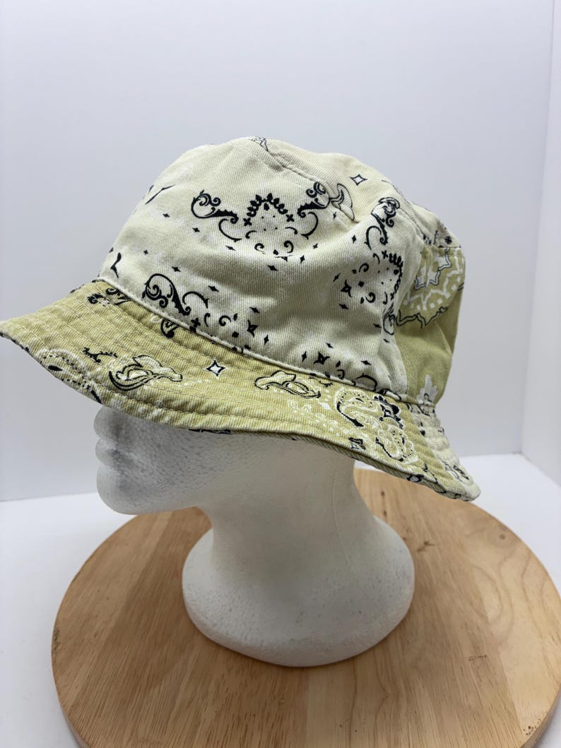 May include: A light green and cream-colored bucket hat with a paisley pattern. The hat features a wide brim and is displayed on a white mannequin head.