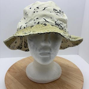 May include: Cream-colored bucket hat with a black and olive green paisley pattern. The hat is displayed on a white mannequin head and a wooden surface. The hat's brim is reversible, showcasing a contrasting design.