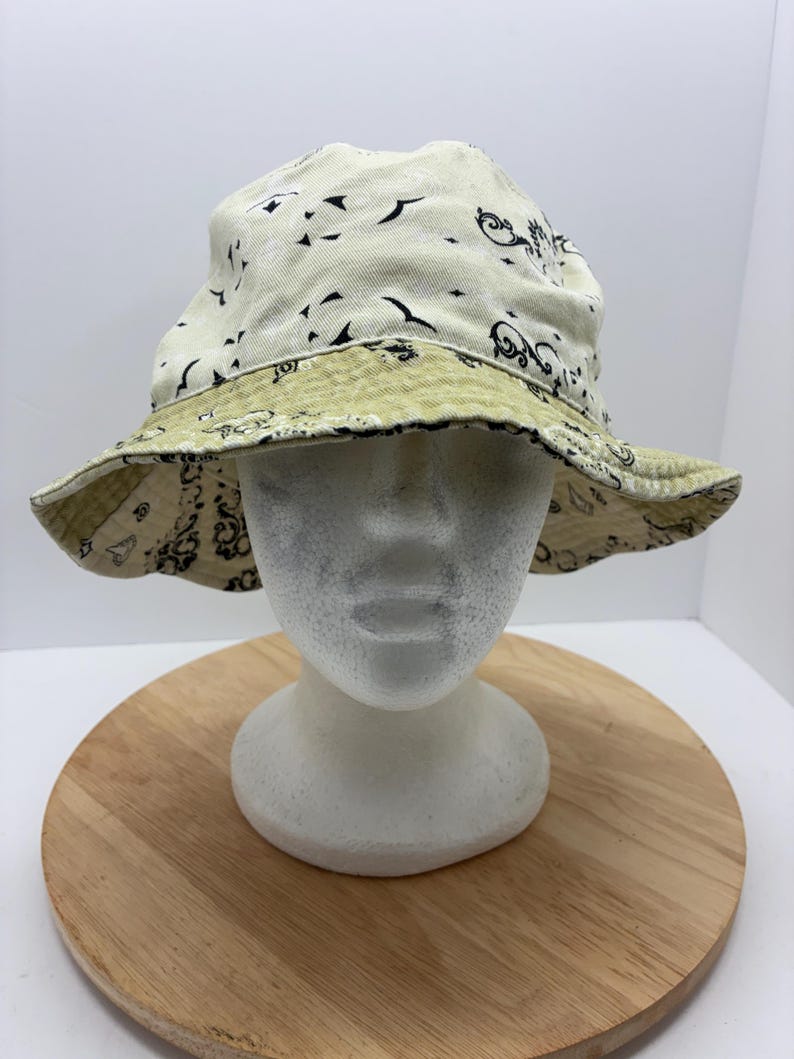 May include: Cream-colored bucket hat with a black and olive green paisley pattern. The hat is displayed on a white mannequin head and a wooden surface. The hat's brim is reversible, showcasing a contrasting design.