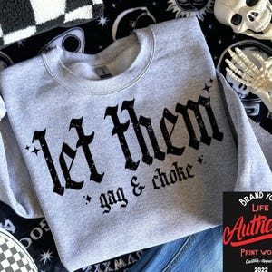 May include: A light gray sweatshirt with the text "Let Them gag & choke" in a black gothic font. The sweatshirt has a crew neck and long sleeves. The image also includes a black and white checkered pattern and a skeleton.