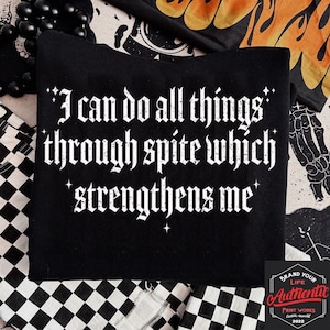 May include: Black sweatshirt featuring the quote "I can do all things through spite which strengthens me" in white gothic lettering. The garment is displayed on a black and white checkered surface, with a beaded necklace and decorative fabric in the background.