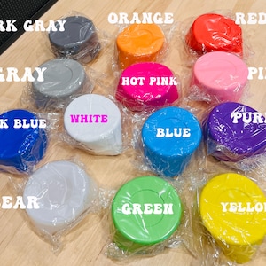 May include: A collection of twelve circular plastic lids in various colors, each individually wrapped in clear plastic. The colors include dark gray, orange, red, gray, hot pink, pink, dark blue, white, blue, purple, clear, green, and yellow.