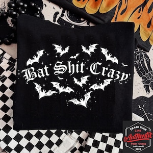 May include: Black sweatshirt with the phrase "Bat Shit Crazy" in a gothic font, surrounded by white bat illustrations. The design includes small stars and is set against a black background. The sweatshirt is part of a Halloween or gothic-themed collection.