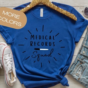 May include: A royal blue t-shirt with the text "Medical Records Squad" in black lettering with a black and white marker graphic. The shirt is on a wooden surface with a gold hanger, white trainers, and blue denim shorts.