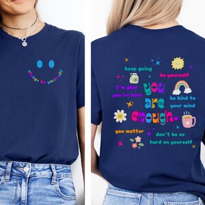 Könnte beinhalten: Marineblaues T-Shirt mit einem Smiley und dem Text "always be yourself" auf der Vorderseite. Auf der Rückseite des Shirts stehen die Worte "you are enough" und andere positive Affirmationen. Das Shirt wird mit einer blauen Jeans getragen.