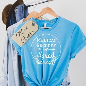 May include: A light blue t-shirt with a white circle graphic that says "Medical Records Squad" with an arrow pointing to the right. The shirt is hanging on a hanger with a brown tag that says "More Colors".