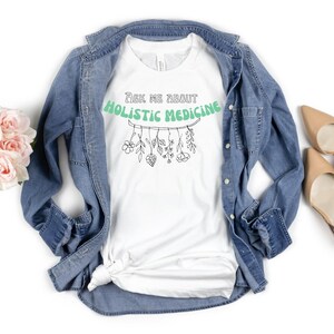 May include: White t-shirt with the text "Ask Me About Holistic Medicine" in a green banner. Below the text is a line drawing of herbs and flowers. The shirt is paired with a denim jacket, nude heels, and a bouquet of pink roses.