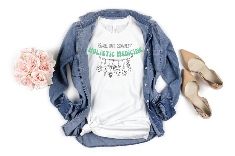 May include: White t-shirt with the text "Ask Me About Holistic Medicine" in a green banner. Below the text is a line drawing of herbs and flowers. The shirt is paired with a denim jacket, nude heels, and a bouquet of pink roses.