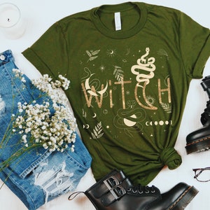 Puede incluir: Una camiseta verde con un estampado gr&aacute;fico dorado de la palabra "WITCH" rodeada de elementos celestiales e inspirados en la naturaleza, como una serpiente, lunas, estrellas y hojas.