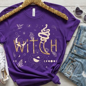 Puede incluir: Camiseta morada con un dise&ntilde;o de purpurina dorada que dice "WITCH" con un gr&aacute;fico de serpiente y luna.