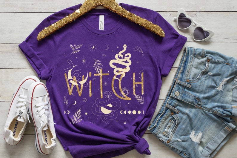 Puede incluir: Camiseta morada con un dise&ntilde;o de purpurina dorada que dice "WITCH" con un gr&aacute;fico de serpiente y luna.