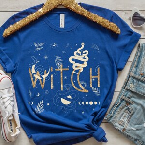 Puede incluir: Una camiseta azul real con un dise&ntilde;o de purpurina dorada de la palabra "WITCH" rodeada de lunas, estrellas y serpientes.