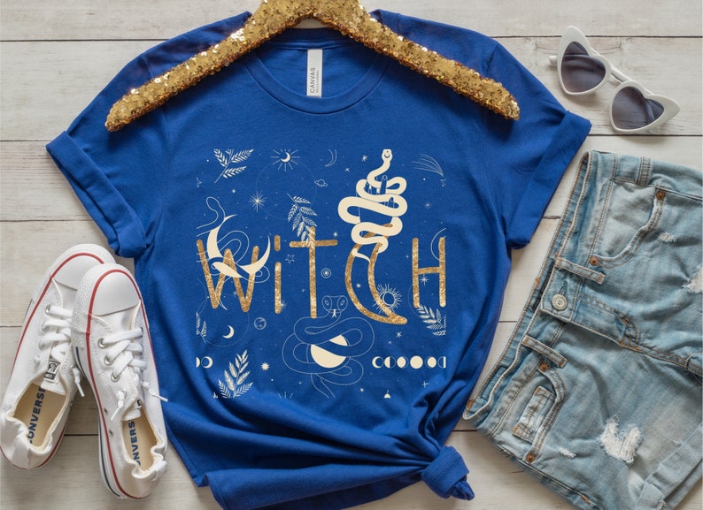 Puede incluir: Una camiseta azul real con un dise&ntilde;o de purpurina dorada de la palabra "WITCH" rodeada de lunas, estrellas y serpientes.