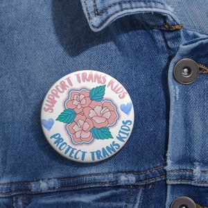 May include: A white button with pink flowers and blue hearts. The text on the button reads "Support Trans Kids Protect Trans Kids".