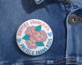 May include: A white button with pink flowers and blue hearts. The text on the button reads "Support Trans Kids Protect Trans Kids".