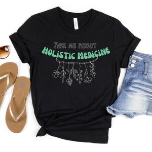 May include: Black t-shirt with the text "Ask me about Holistic Medicine" in a retro font. The shirt features a botanical illustration of hanging plants. Sunglasses, gold flip-flops, and denim shorts are also in the image.