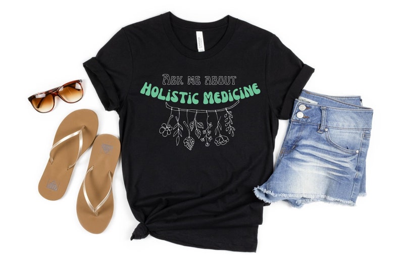 May include: Black t-shirt with the text "Ask me about Holistic Medicine" in a retro font. The shirt features a botanical illustration of hanging plants. Sunglasses, gold flip-flops, and denim shorts are also in the image.