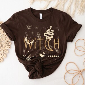 Puede incluir: Una camiseta marr&oacute;n con un dise&ntilde;o gr&aacute;fico dorado que dice "WITCH" con una serpiente, una luna y estrellas.