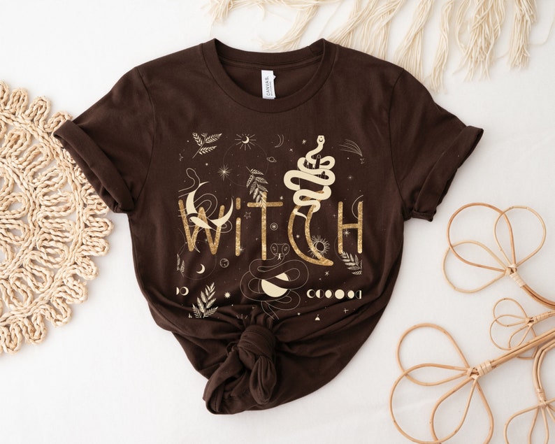 Puede incluir: Una camiseta marr&oacute;n con un dise&ntilde;o gr&aacute;fico dorado que dice "WITCH" con una serpiente, una luna y estrellas.