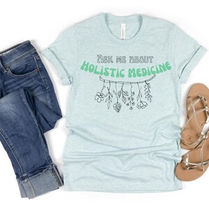 May include: Light blue t-shirt with rolled sleeves, jeans, and sandals. The shirt features the text "Ask Me About Holistic Medicine" in green, with a botanical illustration. The jeans are blue denim. The sandals are gold.