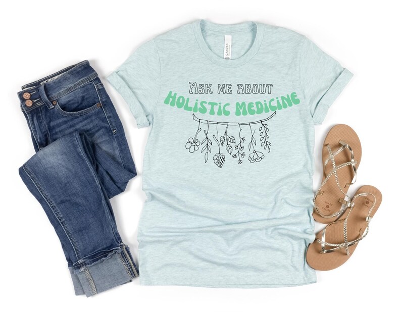 May include: Light blue t-shirt with rolled sleeves, jeans, and sandals. The shirt features the text "Ask Me About Holistic Medicine" in green, with a botanical illustration. The jeans are blue denim. The sandals are gold.